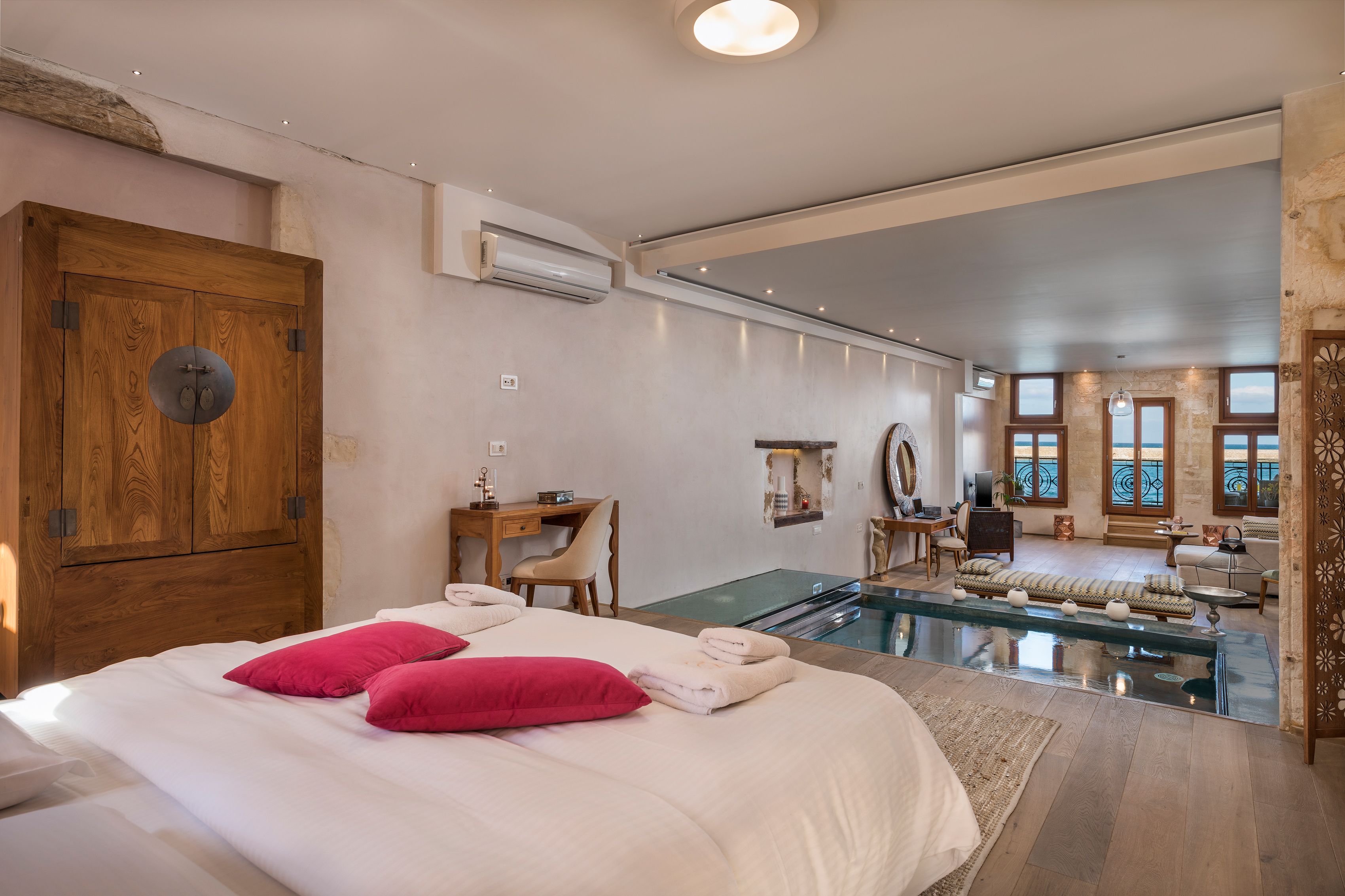 A spacious bedroom with a large bed, a desk, and a view of a living area with a pool.