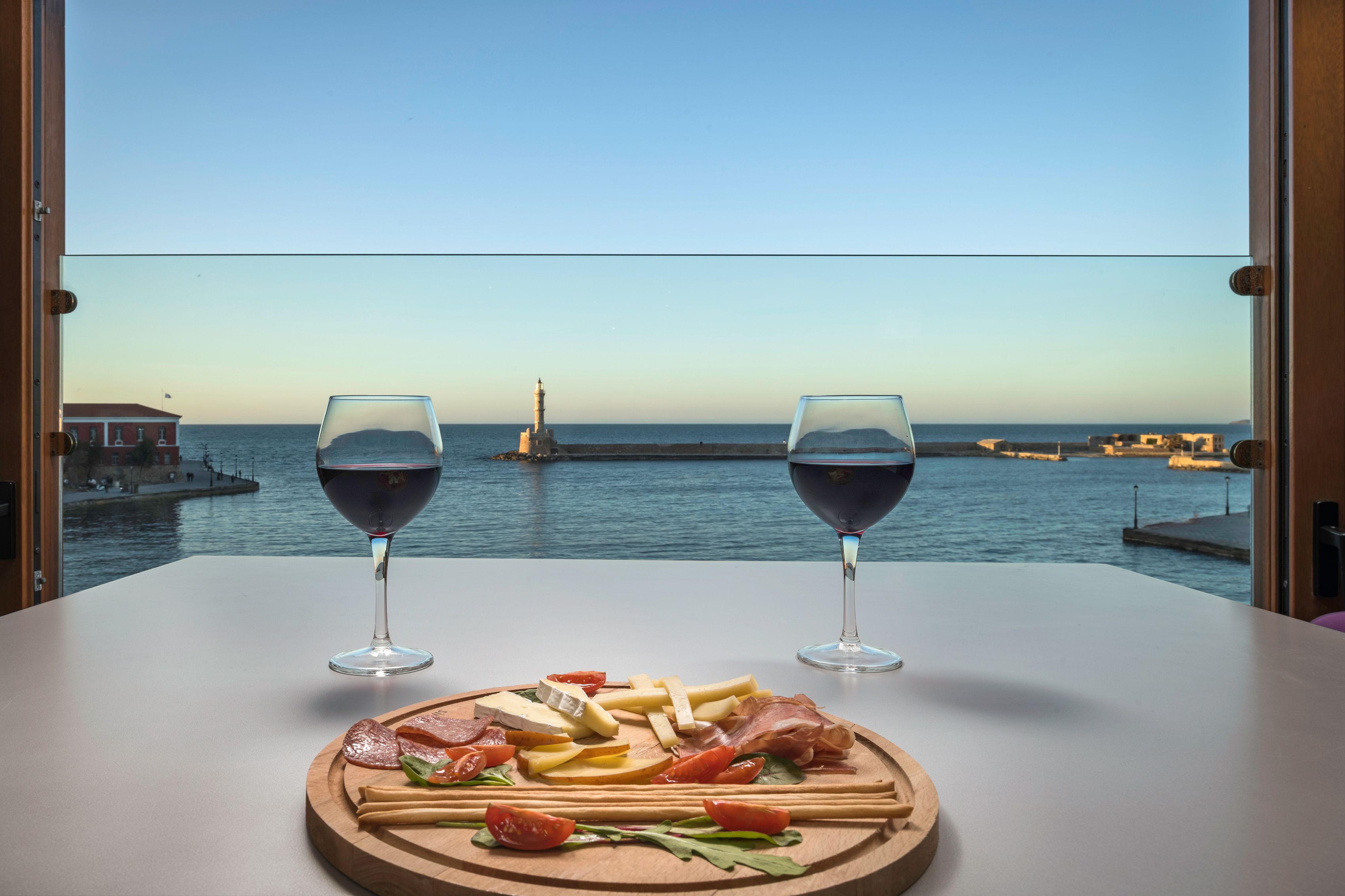 Una mesa con una tabla de embutidos y dos copas de vino tinto, con vista a un puerto con barcos y un faro.