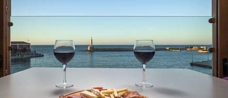 Una mesa con una tabla de embutidos y dos copas de vino tinto, con vista a un puerto con barcos y un faro.