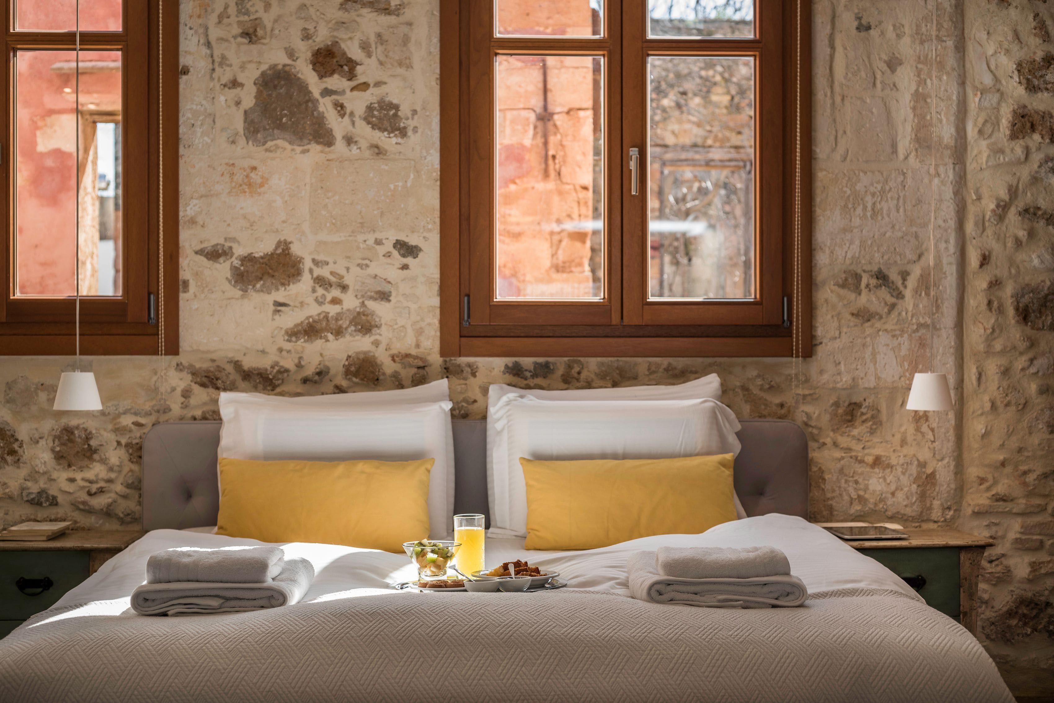 A hotel room with a stone wall, two windows, a bed with white and yellow pillows, a bedside table with a lamp, and a breakfast tray on the bed.