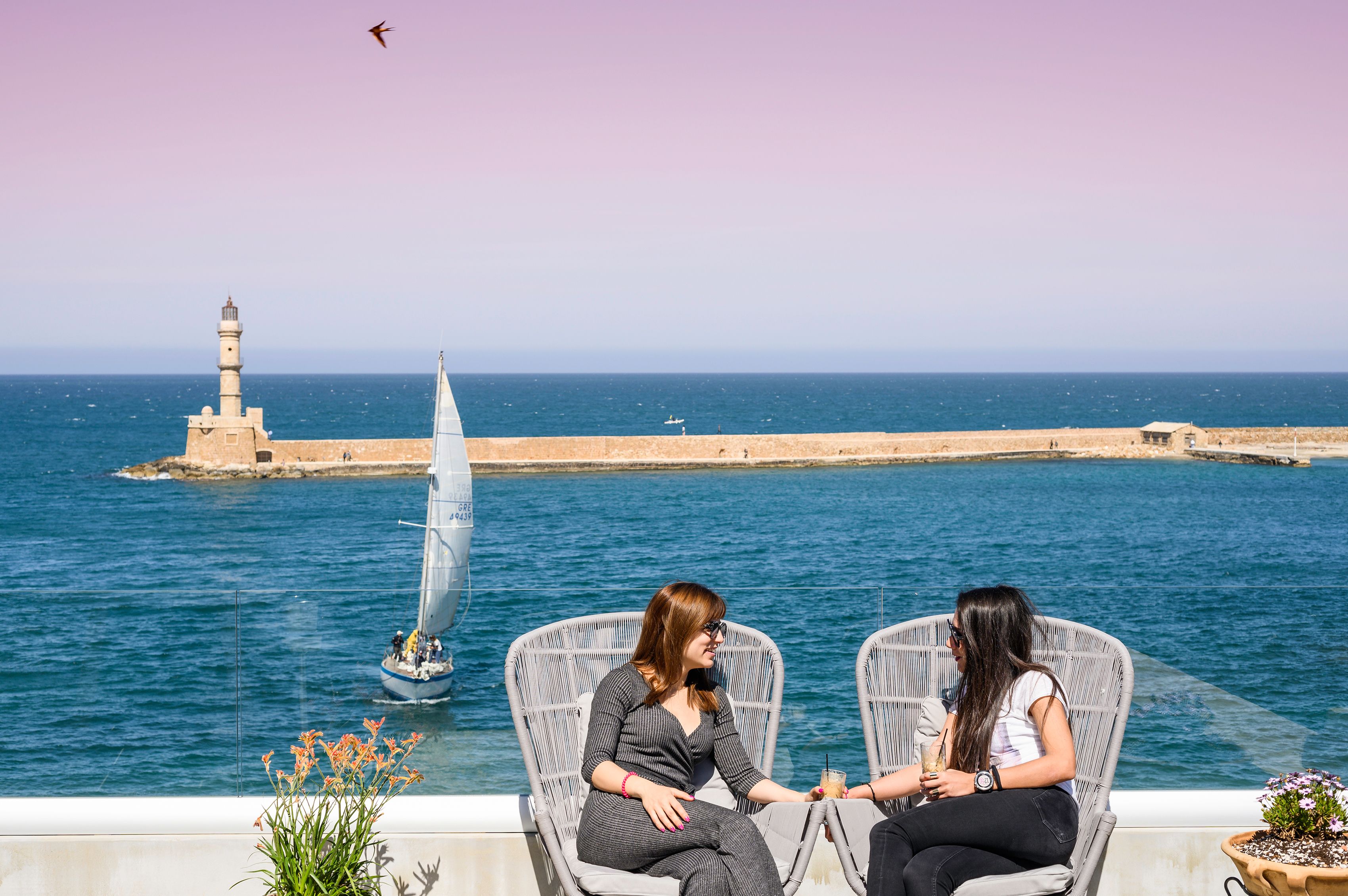 Two people seated on wicker chairs overlooking a body of water with a lighthouse in the background.