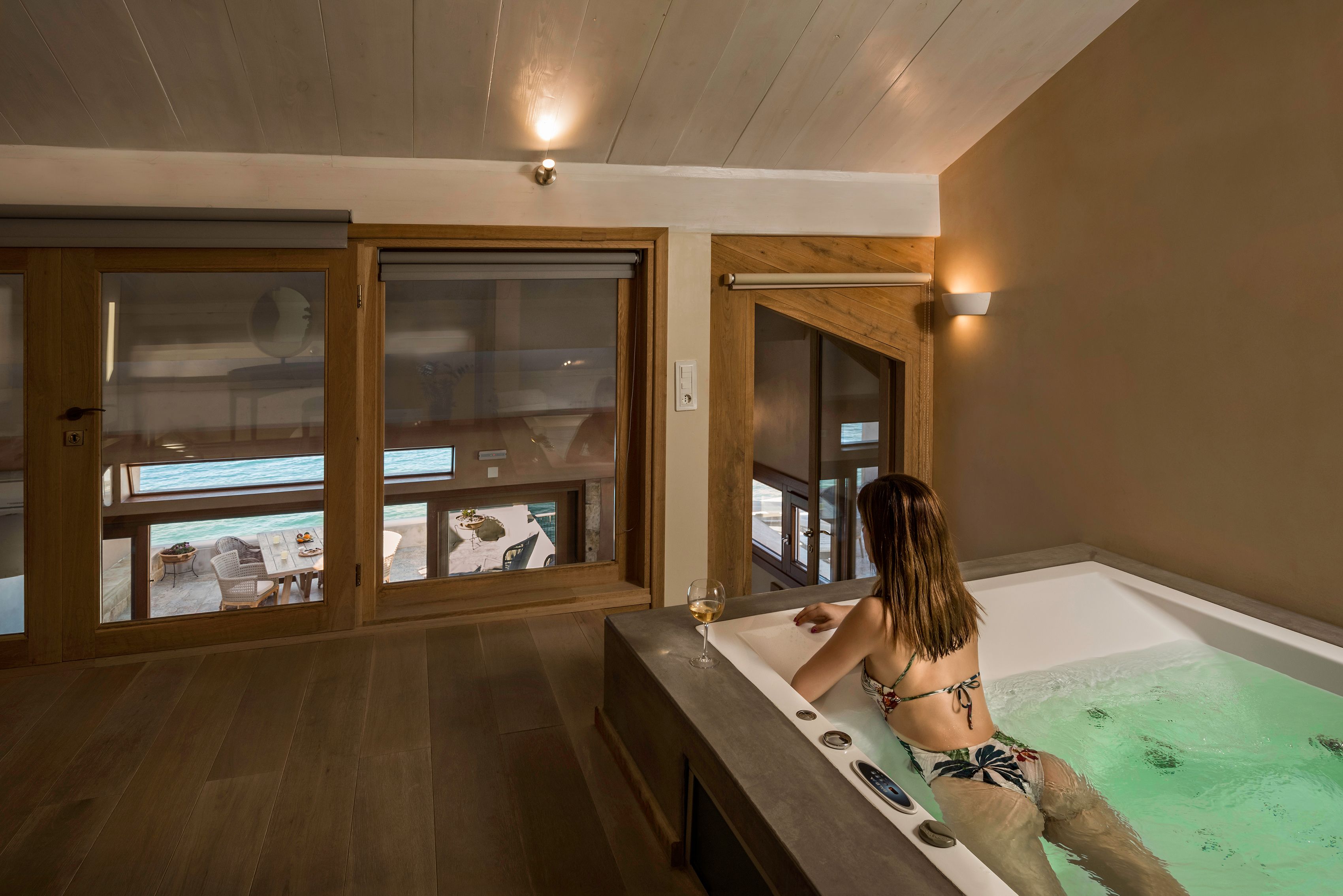 A woman relaxing in a bathtub with a glass of wine, wooden floors, and large windows overlooking a balcony area.
