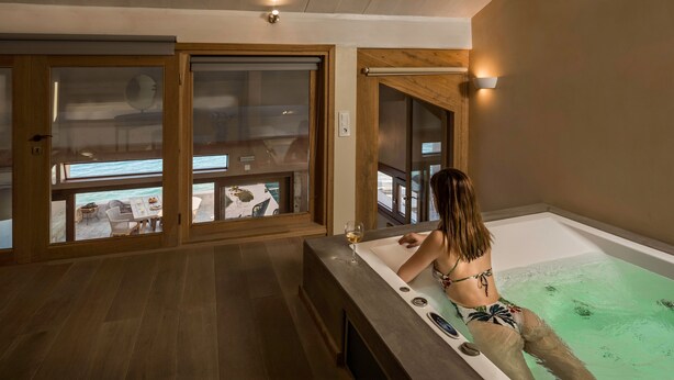A woman relaxing in a bathtub with a glass of wine, wooden floors, and large windows overlooking a balcony area. - Vranas Residenza Signature Suites (Chania)