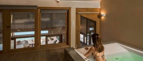 A woman relaxing in a bathtub with a glass of wine, wooden floors, and large windows overlooking a balcony area.