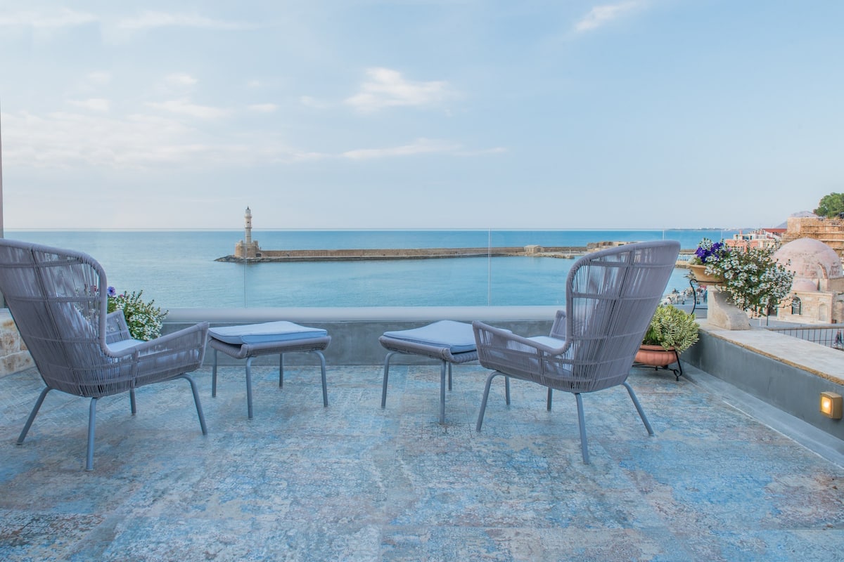 A terrace with two wicker chairs and a small table, overlooking a calm sea and a lighthouse.