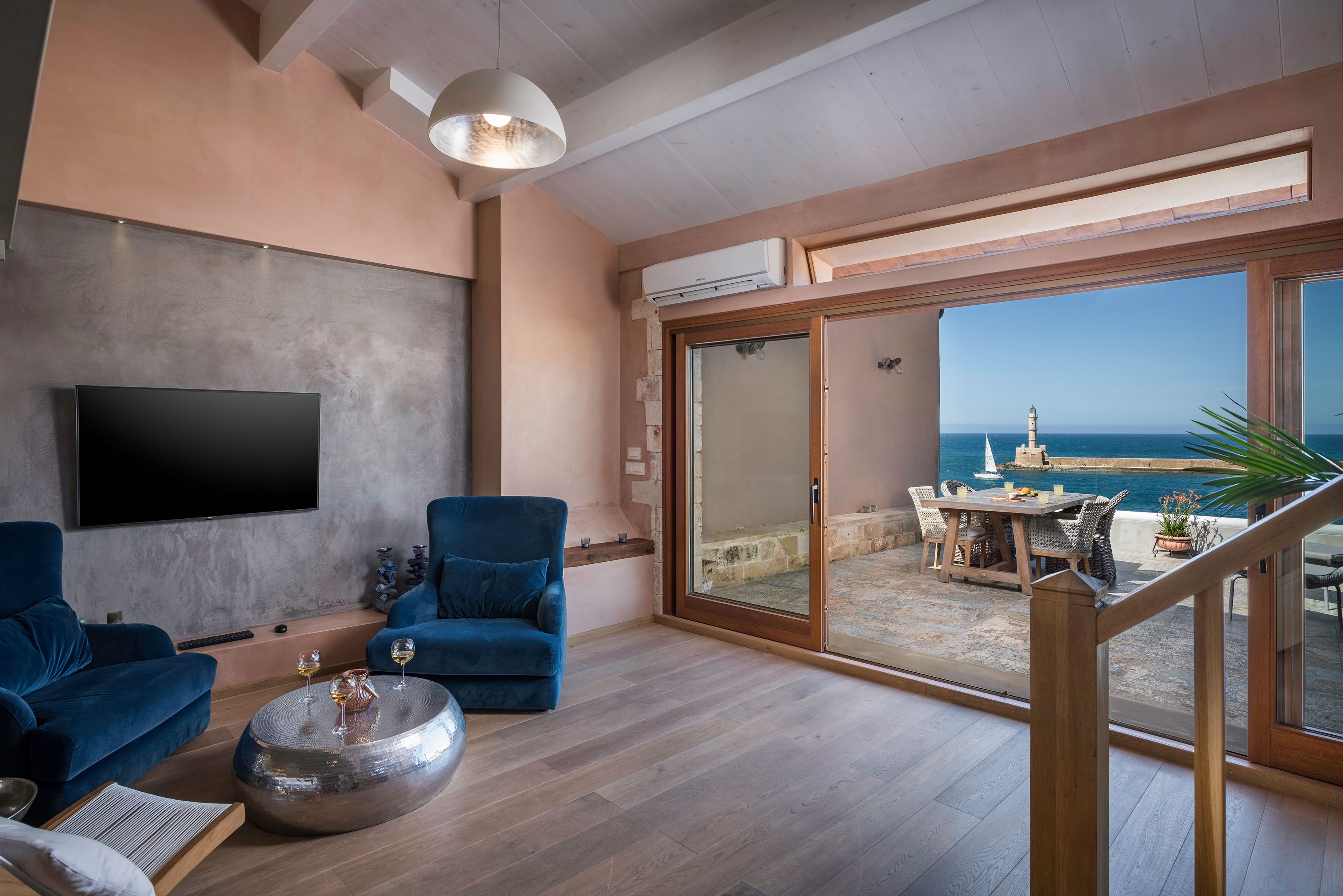 A modern living room with a flat-screen TV, blue sofas, a round coffee table, and a view of the sea through large sliding glass doors.