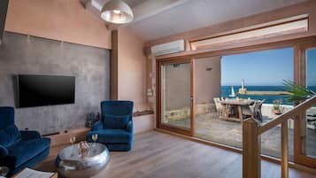 A modern living room with a flat-screen TV, blue sofas, a round coffee table, and a view of the sea through large sliding glass doors.