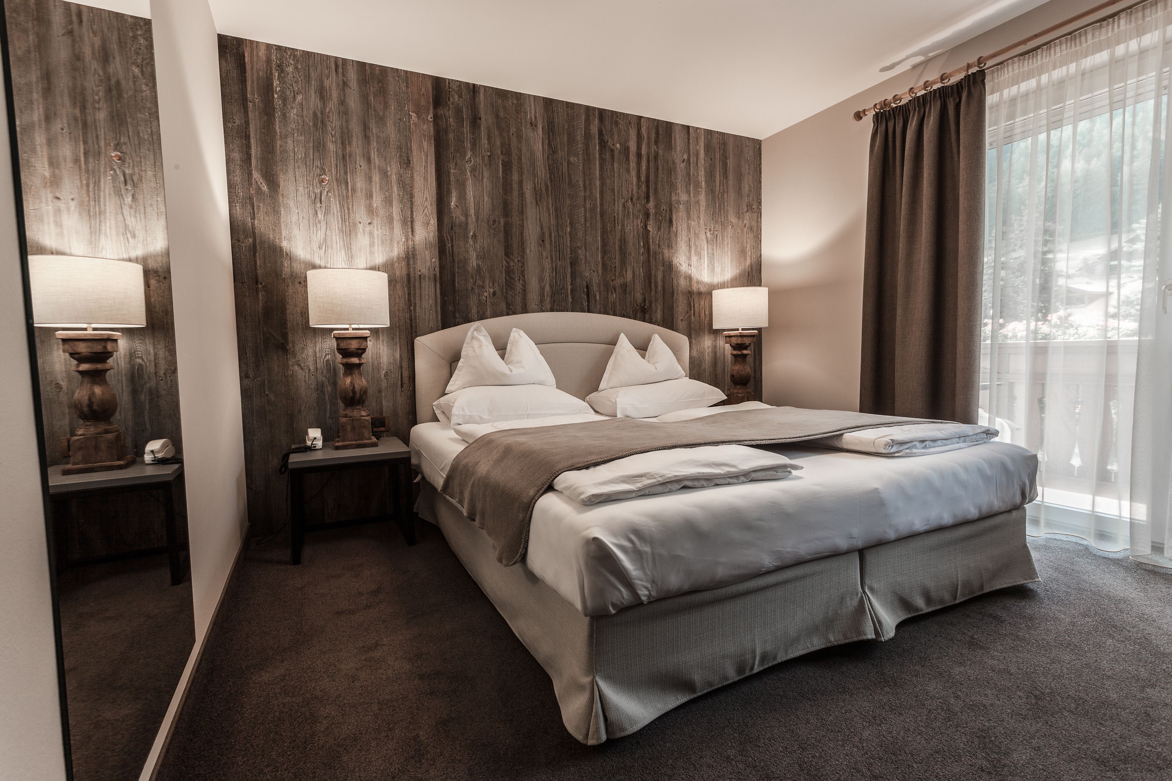 Une chambre d’hôtel moderne avec un grand lit, deux tables de chevet avec des lampes, un mur décoratif en bois et une grande fenêtre avec des rideaux.