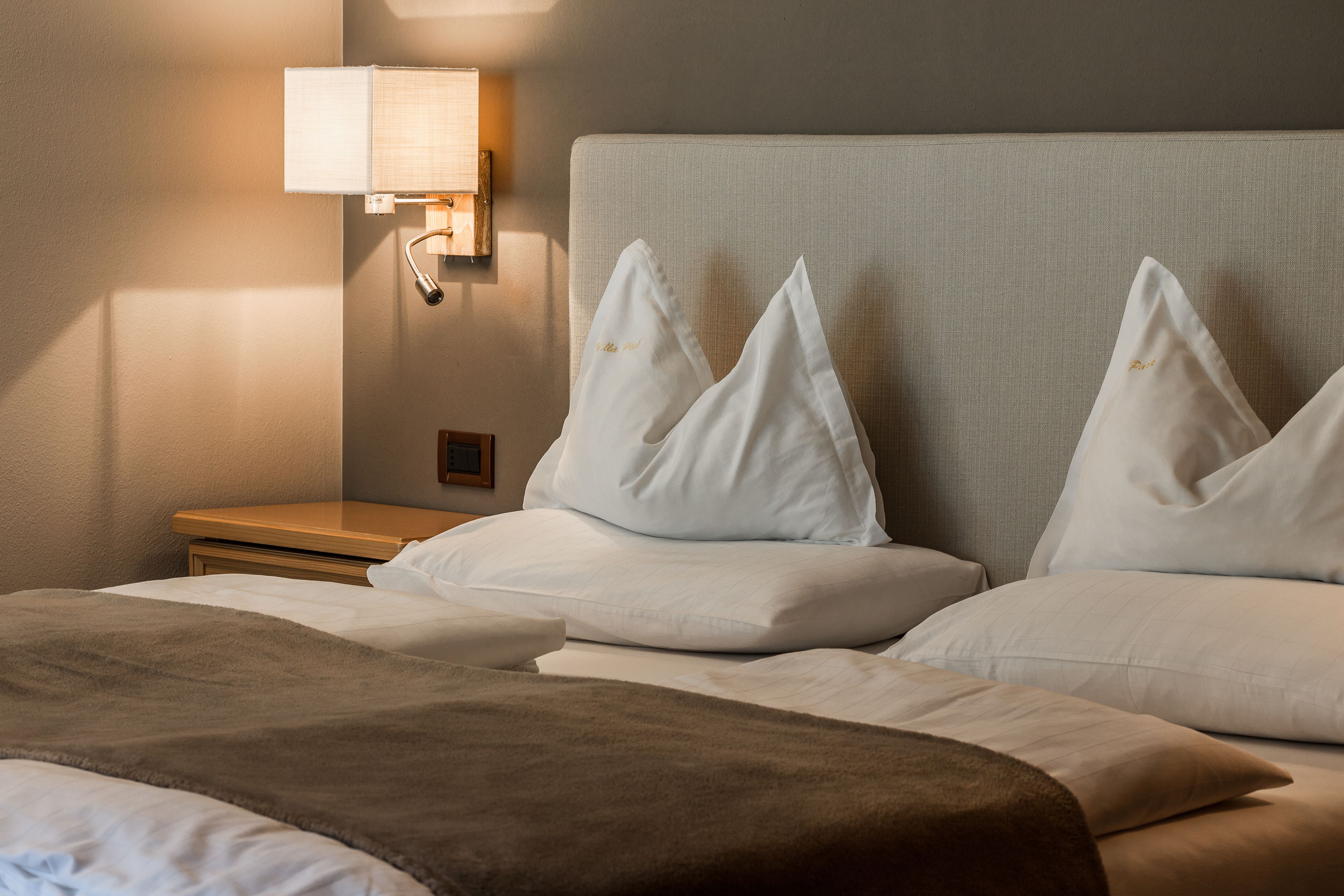Una cama bien tendida con almohadas blancas y una manta marrón, una lámpara de noche y una mesita de madera.