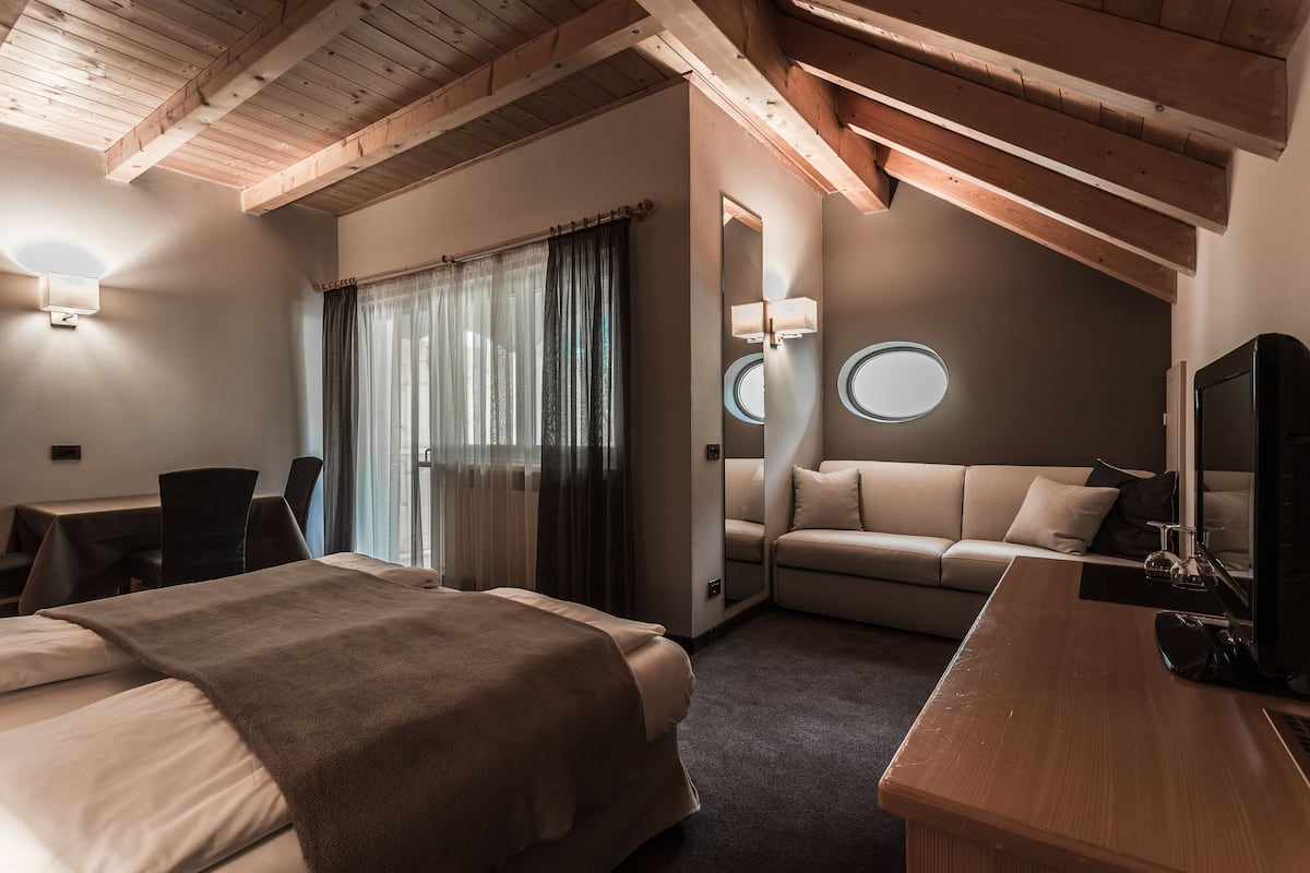 Habitación de hotel con techo de madera, cama con manta marrón, sofá, escritorio con computadora y espejo redondo.