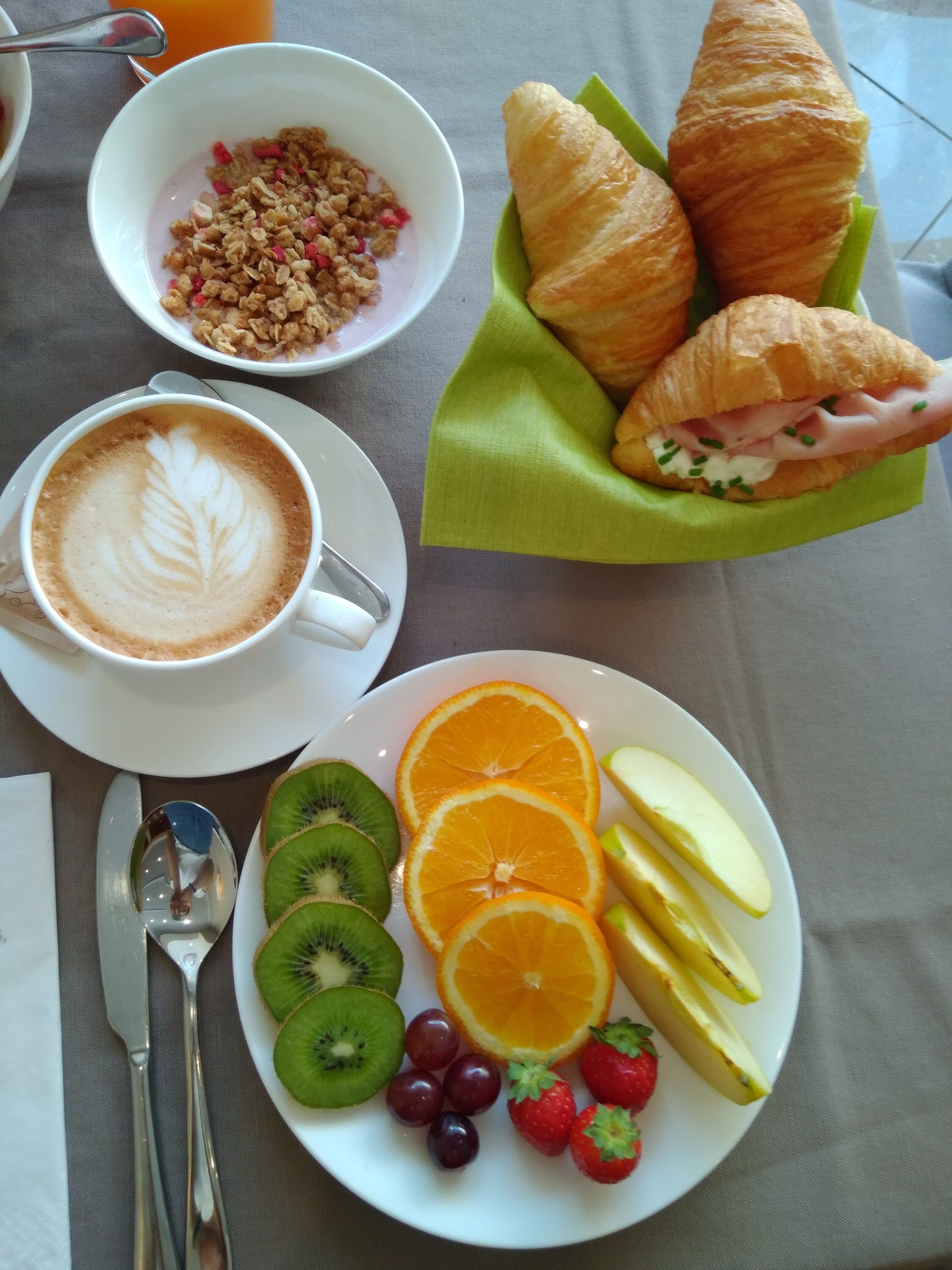 Ein Frühstückstisch mit einem Croissant-Sandwich, Obst, Müsli und einer Kaffeetasse.