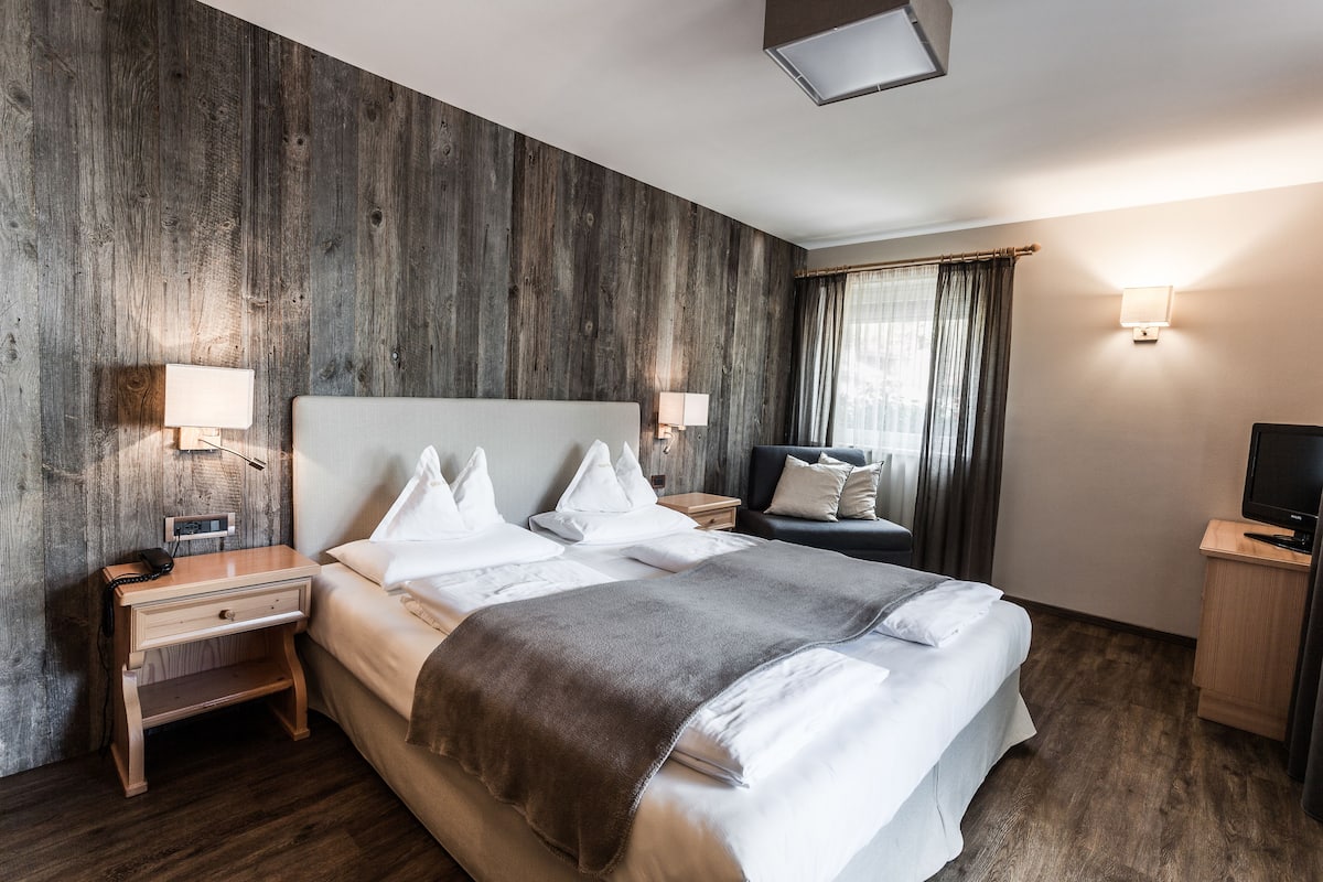 Habitación de hotel con una cama grande, dos mesitas de noche con lámparas, un mueble para televisión y una ventana con cortinas.