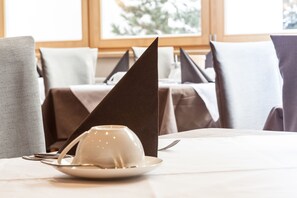 A dining table with a white plate and a cup, set with cutlery, in a room with a window and a plant in the background. - Hotel Garni Villa Park (Ortisei)