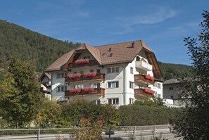 A multi-story hotel with balconies adorned with red flowers, set against a backdrop of a forested mountain. - Hotel Garni Villa Park (Ortisei)