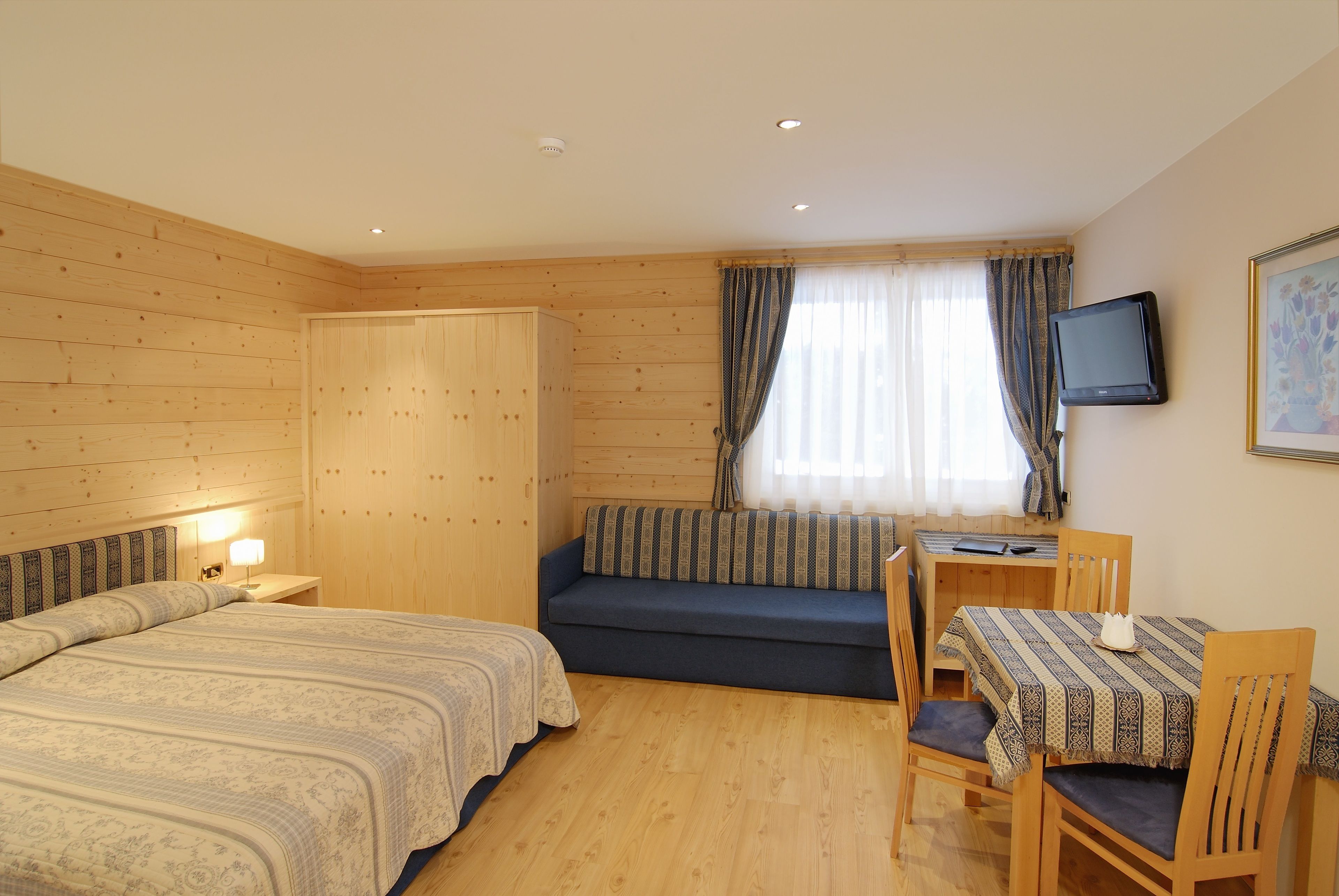 A room with wooden walls, a bed with patterned bedding, a blue sofa, a small table with a TV, and a window with curtains.