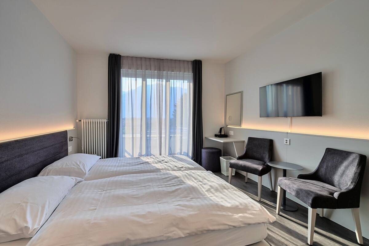 A hotel room with a large bed, two chairs, a small table, a TV mounted on the wall, and a window with curtains.