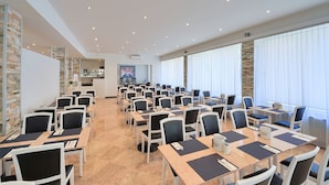 Un salón de conferencias con varias mesas y sillas, una zona de cocina al fondo y ventanas grandes con cortinas.