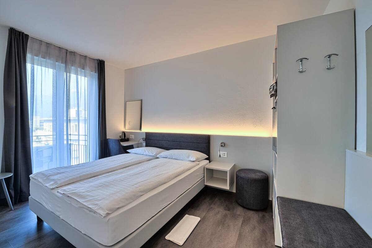 A modern hotel room with a large bed, bedside tables, a small seating area, and a view of the outdoors.