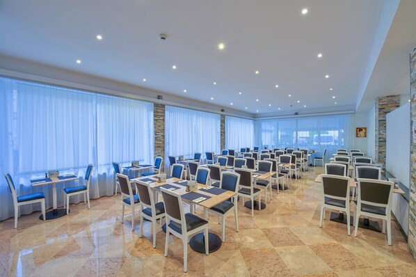 A conference room with multiple tables and chairs, large windows with curtains, and a polished floor.