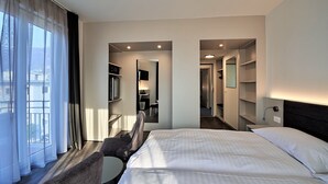 Habitación de hotel con cama, una silla, un escritorio y vistas al exterior a través de cortinas translúcidas.