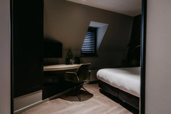 A compact bedroom with a bed, a desk, and a chair. There is a window with blinds, a radiator, and a television on the wall.