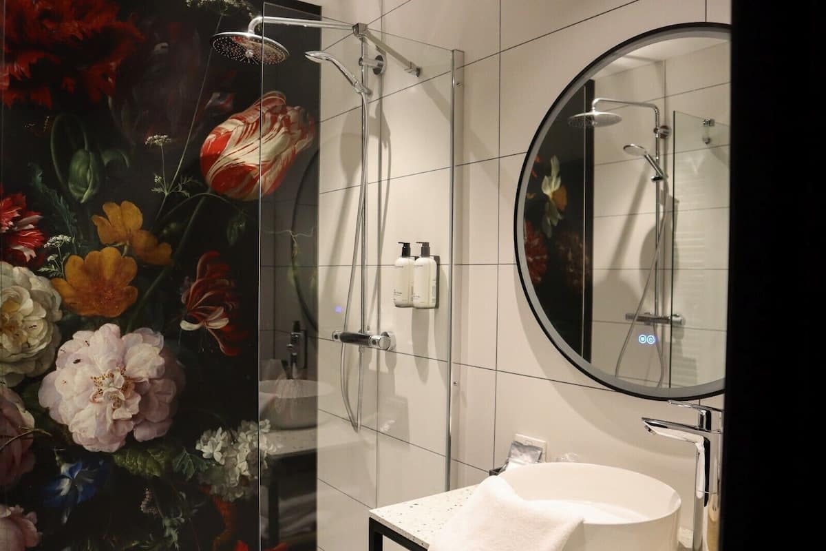 Un baño con cabina de ducha de vidrio, un espejo redondo, un lavabo blanco y un diseño de papel tapiz floral.