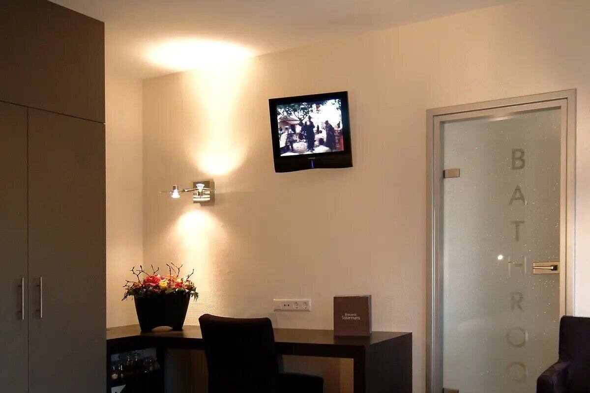 Una habitación de hotel con una cama, un escritorio con computadora, una silla, una planta en maceta, un televisor y una puerta al baño.