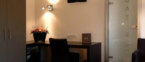 A hotel room with a bed, a desk with a computer, a chair, a potted plant, a television, and a bathroom door.