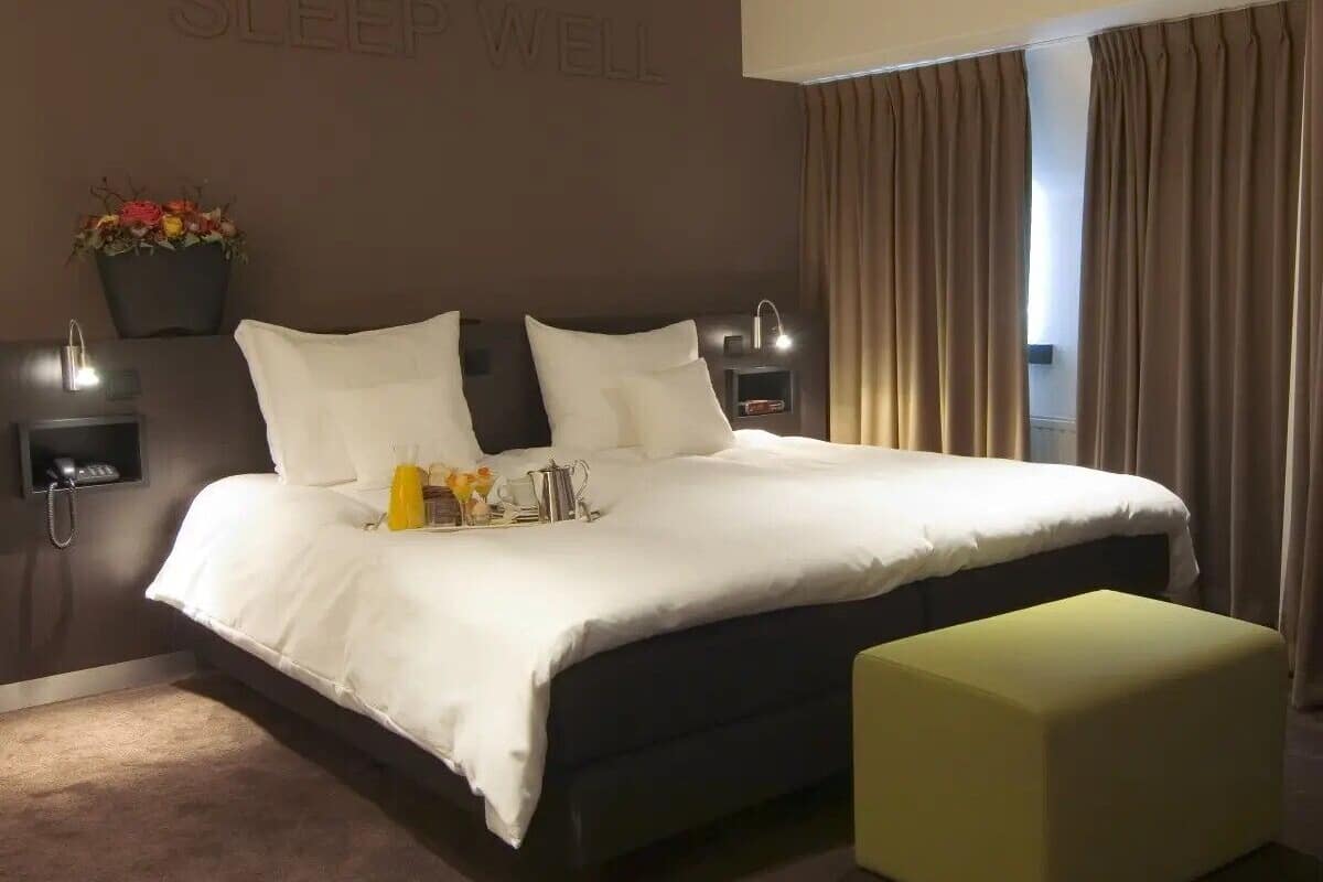 Una habitación de hotel moderna con una cama grande, una mesita de noche con una lámpara, una mesita auxiliar con un teléfono y una luz de pared.
