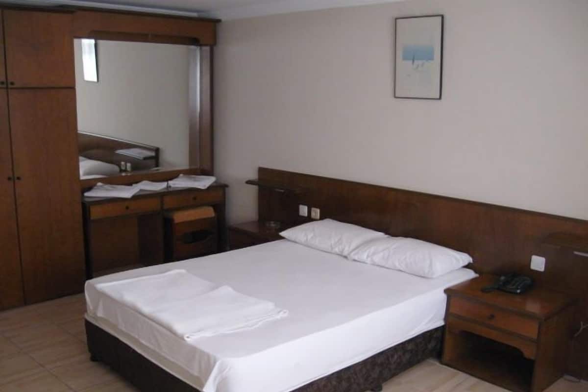 A hotel room with a bed, a wooden wardrobe, a desk with a mirror, and a bedside table with a phone.