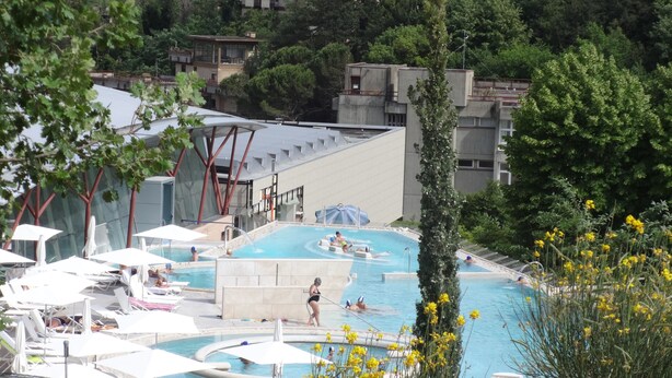 Ett poolområde med flera pooler, solstolar och en byggnad med ett platt tak i bakgrunden.