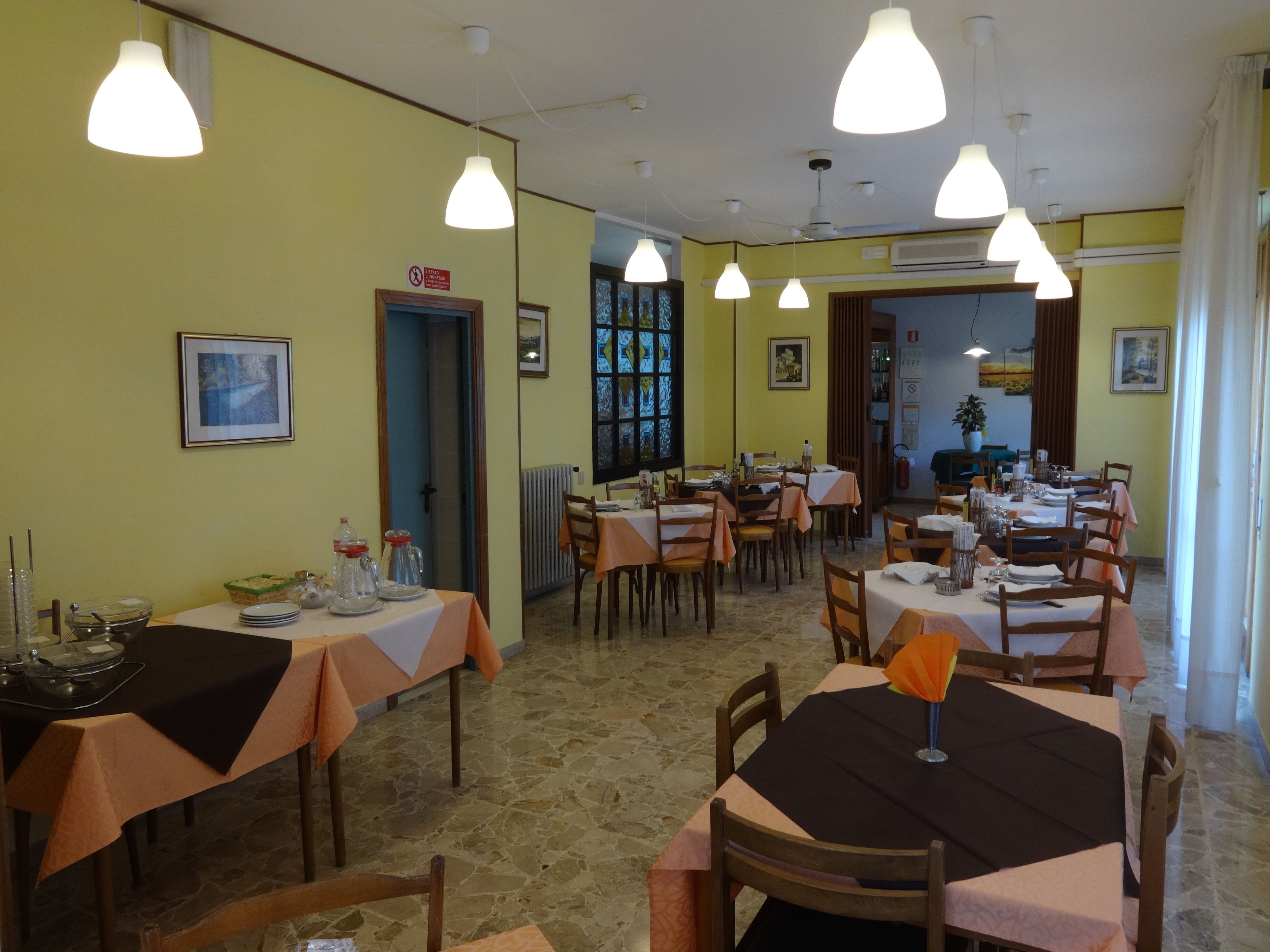 Un comedor con varias mesas, sillas e iluminación colgante. Se observan cuadros en las paredes y una puerta que da a otra habitación.