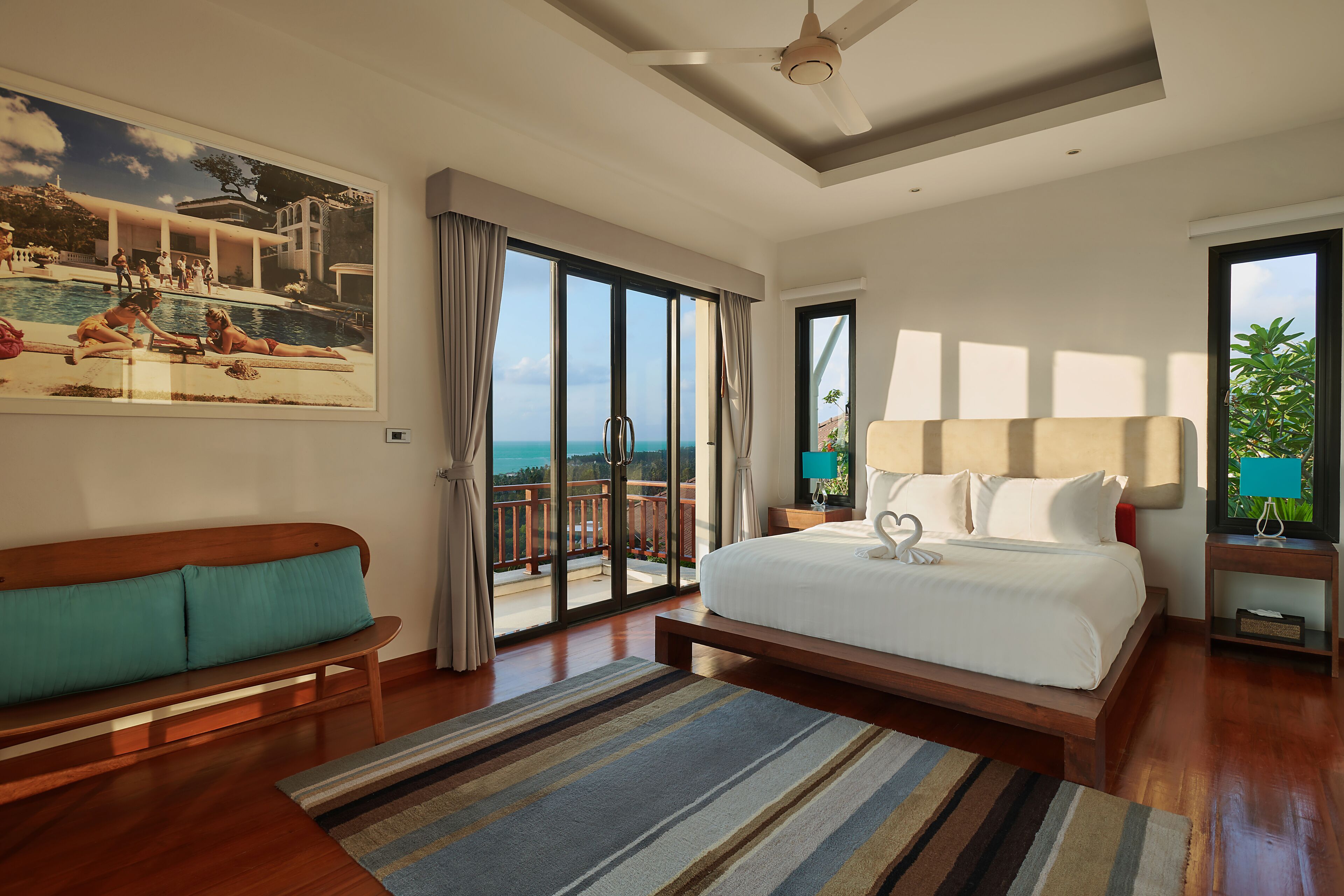 A bedroom with a large bed, a sofa, a view of the ocean, and a painting of a pool scene.