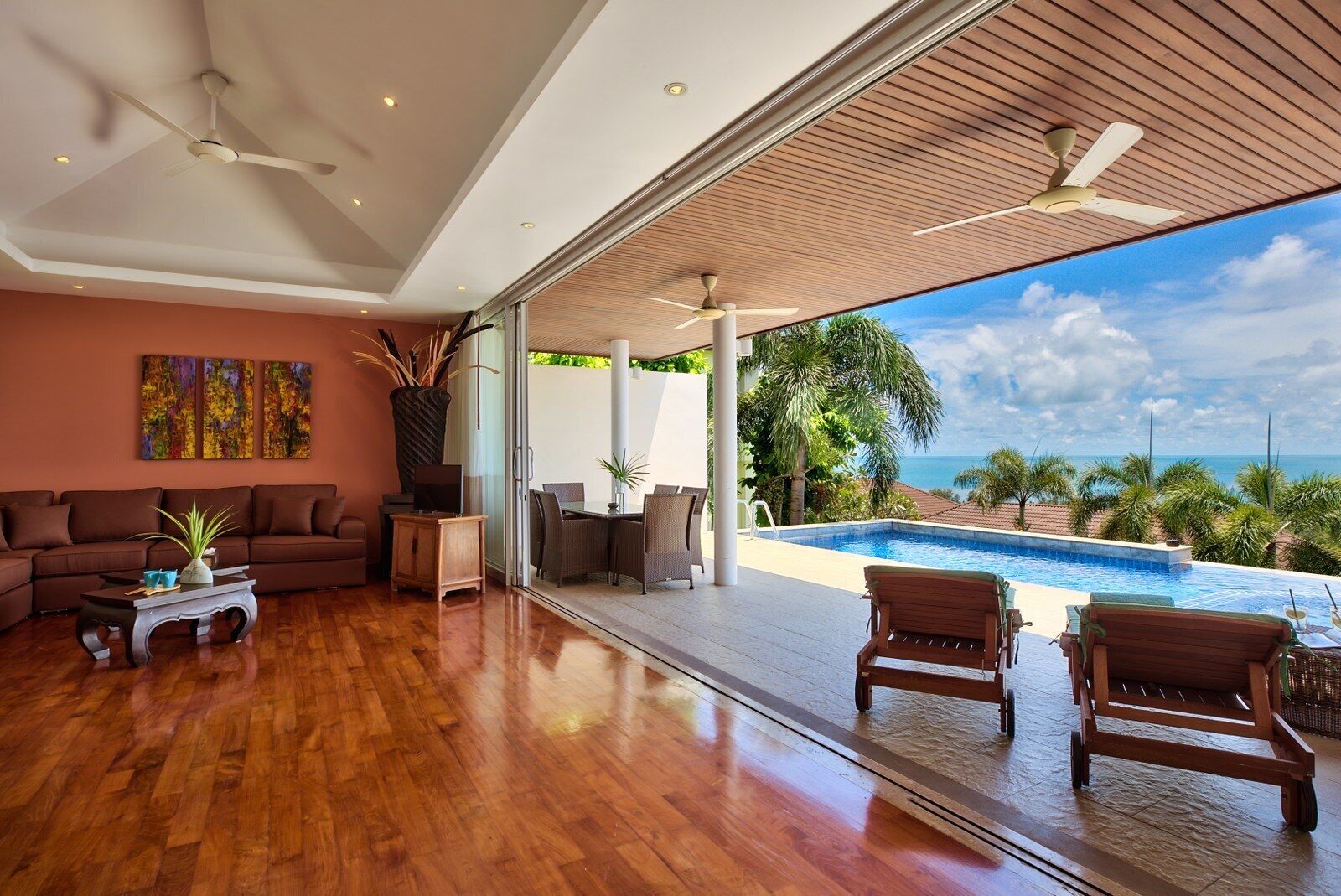 A modern living room with a wooden floor, a large sofa, a coffee table, and a view of a swimming pool and ocean.