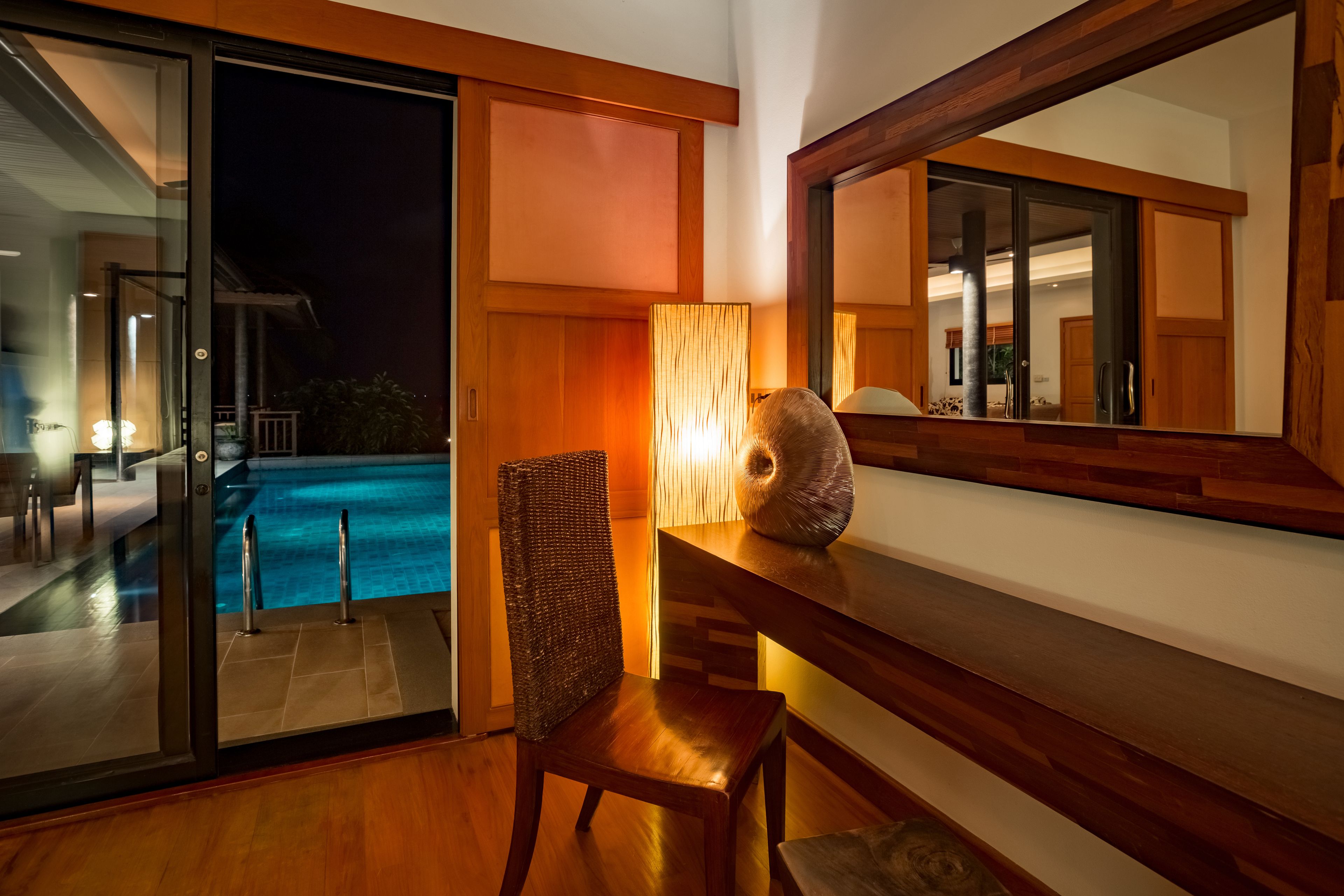A modern interior with a glass door leading to a pool, a wooden chair, and a table with a lamp.