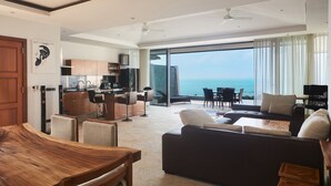 A modern living room with a dark sofa, wooden coffee table, and a dining area with a wooden table and chairs. A kitchen is visible in the background, and there is a large window offering a view of the ocean. - Villa Baan Saitara (Koh Samui)