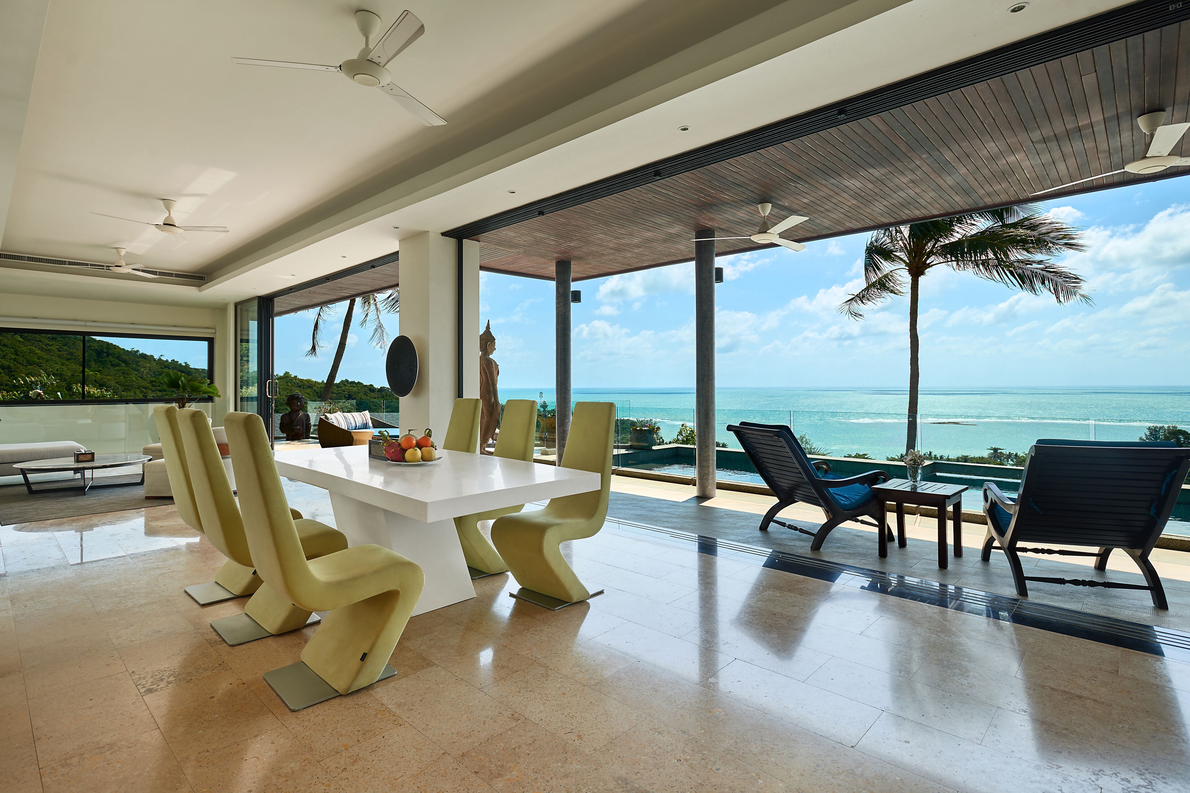 A modern dining area with a large table and chairs, a ceiling fan, and a view of the ocean.