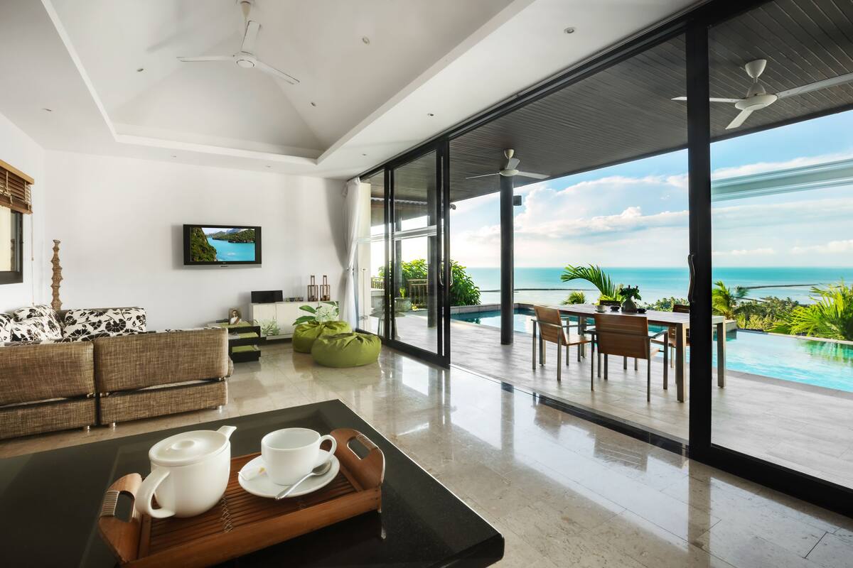 Una sala de estar moderna con un amplio ventanal que da a una zona de piscina, un sofá, una mesa de centro con una bandeja y un televisor en la pared.