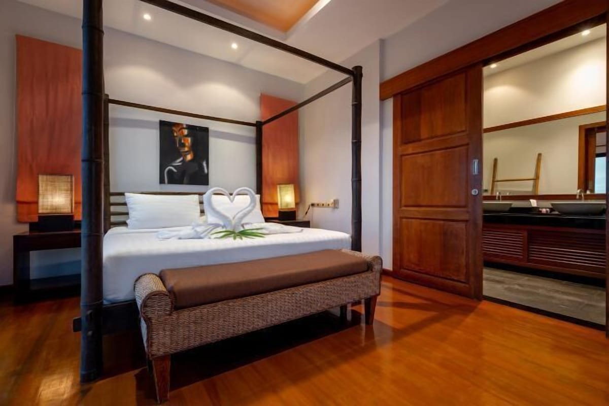 Una cama con dosel, con un arreglo de almohadas con forma de corazón, un banco al pie de la cama y piso de madera.