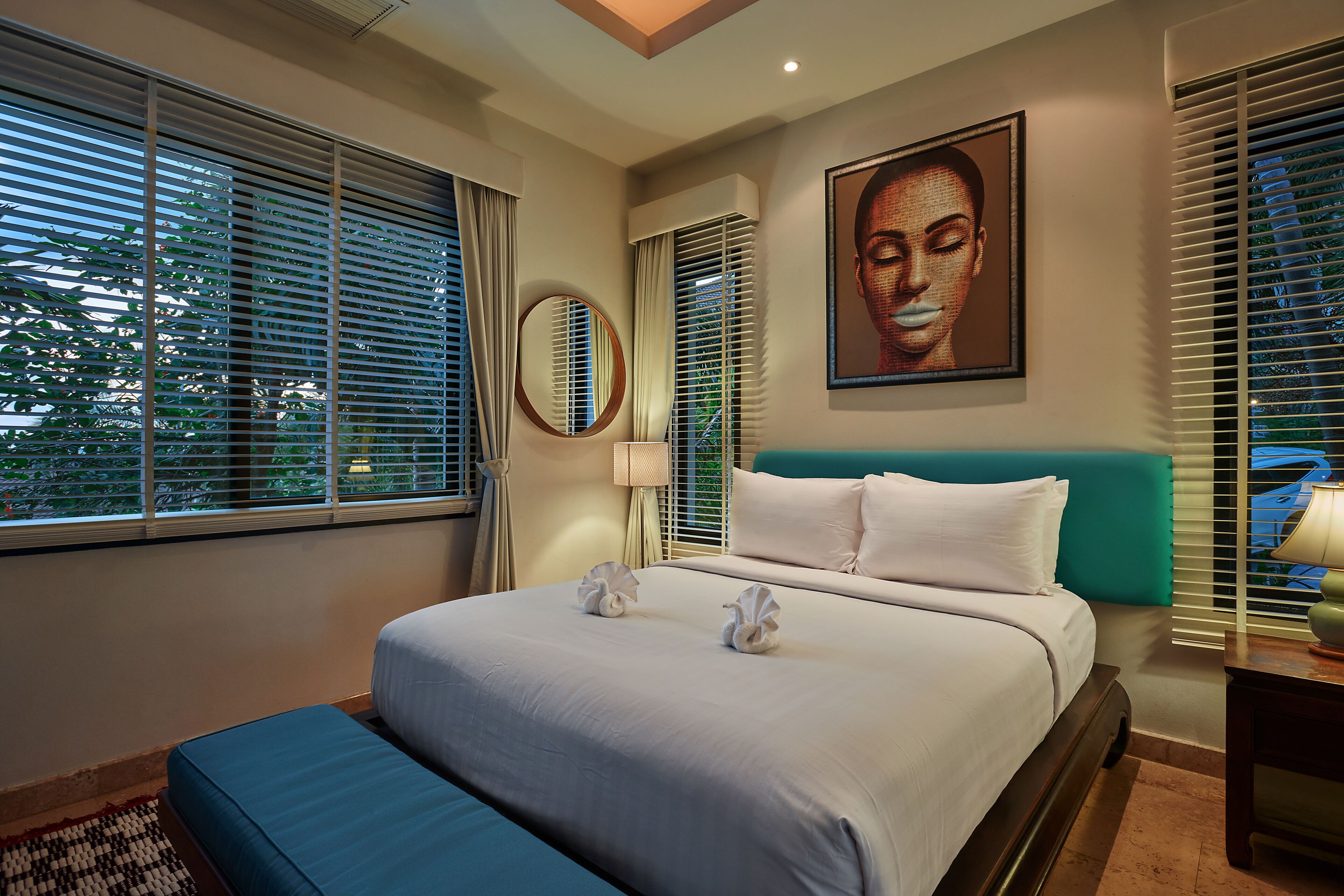 A hotel room with a large bed, a round mirror, a painting on the wall, and a window with blinds.