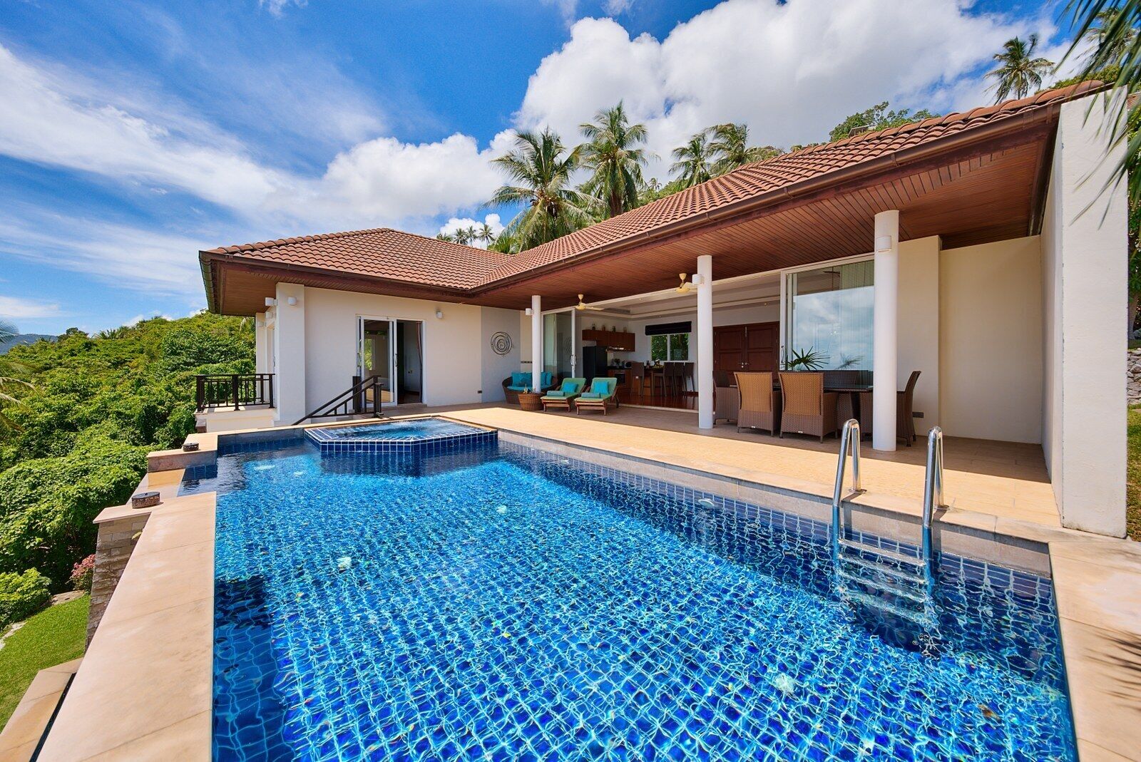 A swimming pool with a tiled deck, a covered outdoor seating area with chairs, and a view of palm trees.