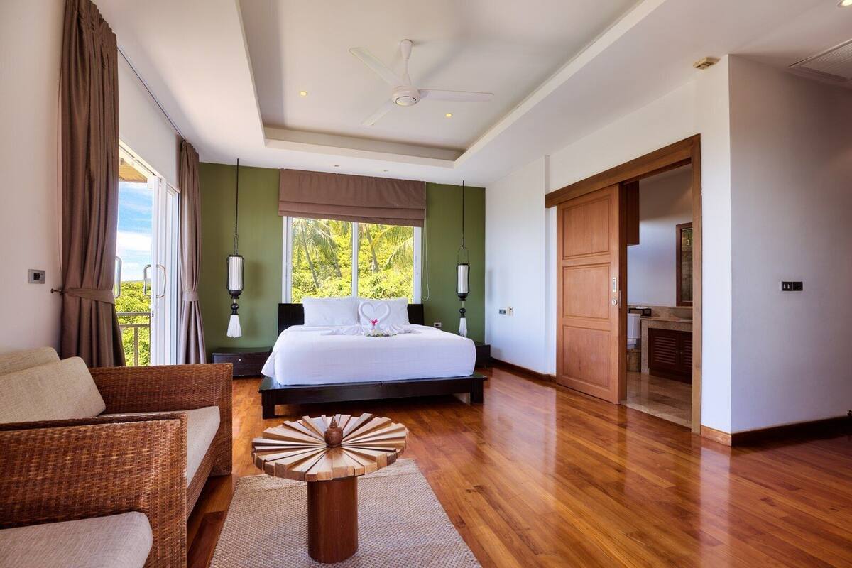 Un dormitorio con cama, ventilador de techo, ventana con cortinas, puerta de madera y sofá de mimbre.