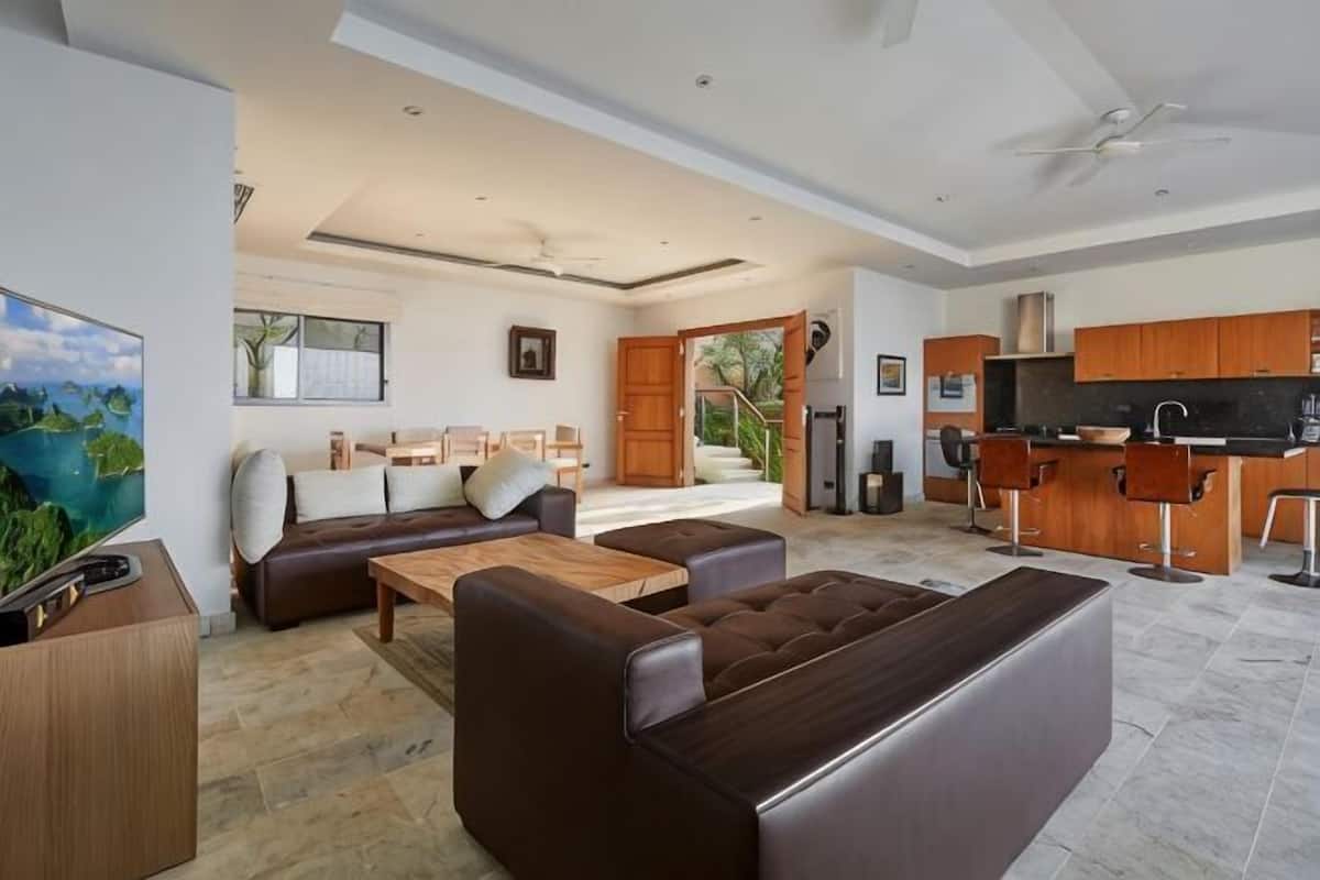 Amplia sala de estar con sofá, mesa de centro y televisor. Se observa una cocina con armarios de madera y una barra.
