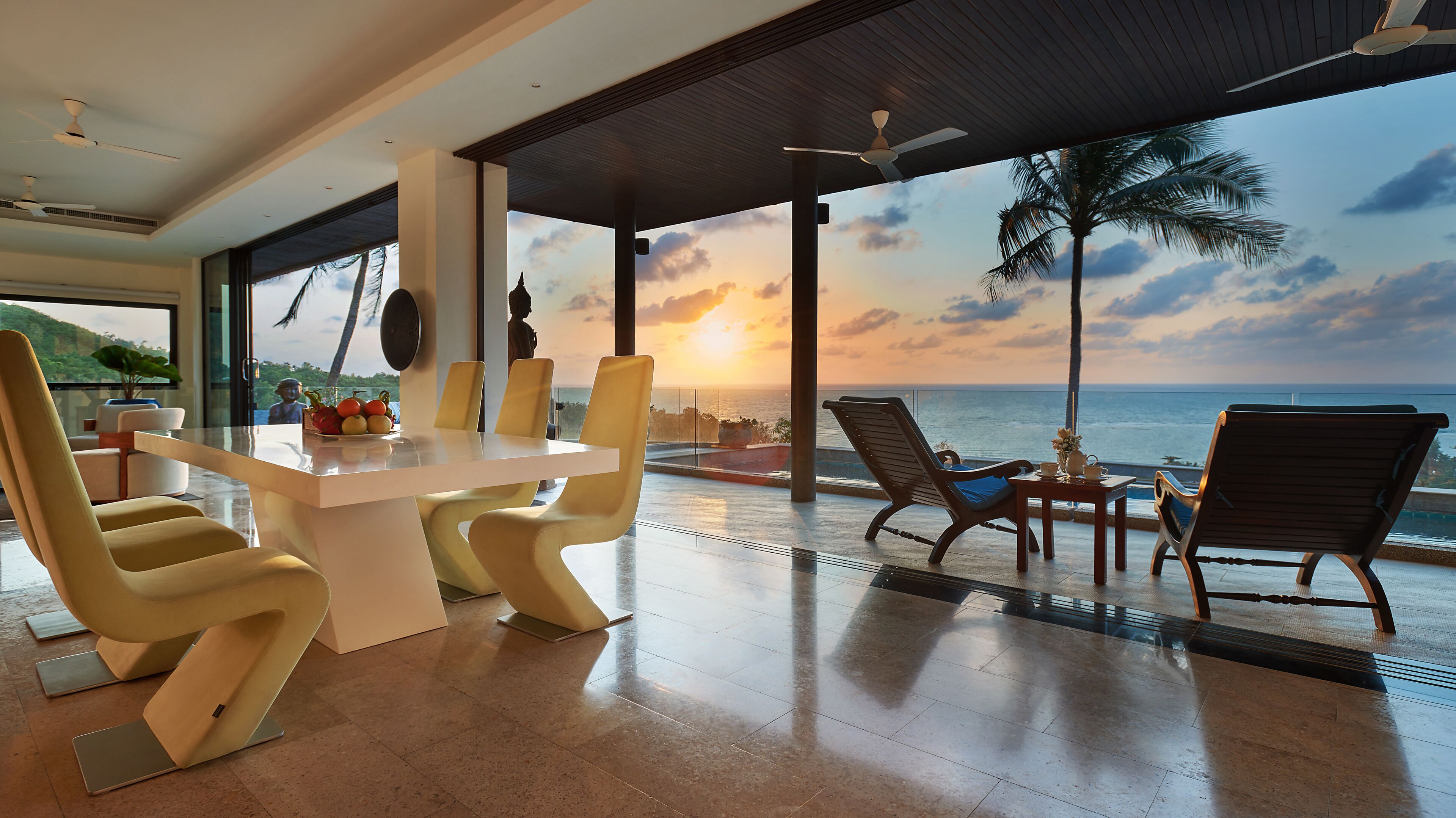 Un espace repas moderne avec vue sur l’océan au coucher du soleil, comprenant une table blanche, des chaises jaunes et une cloison en verre.