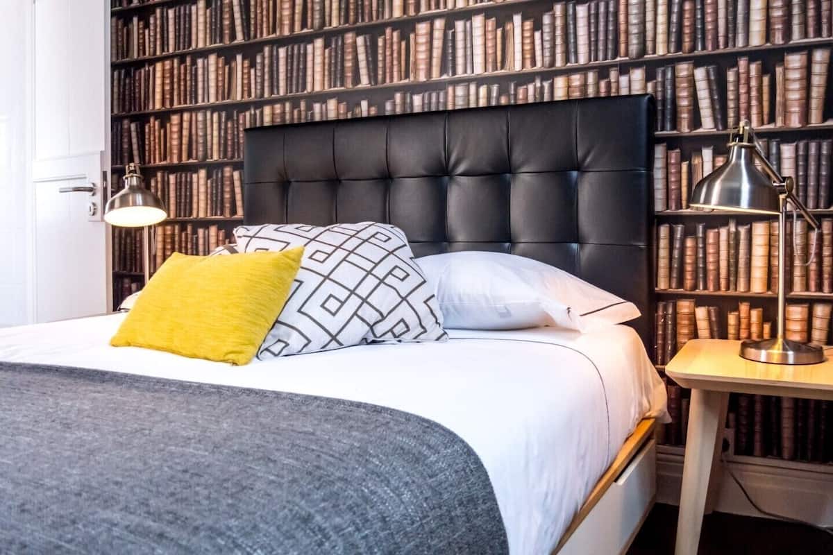 A neatly arranged bedroom with a large bed, a bookshelf filled with books, and a bedside lamp.