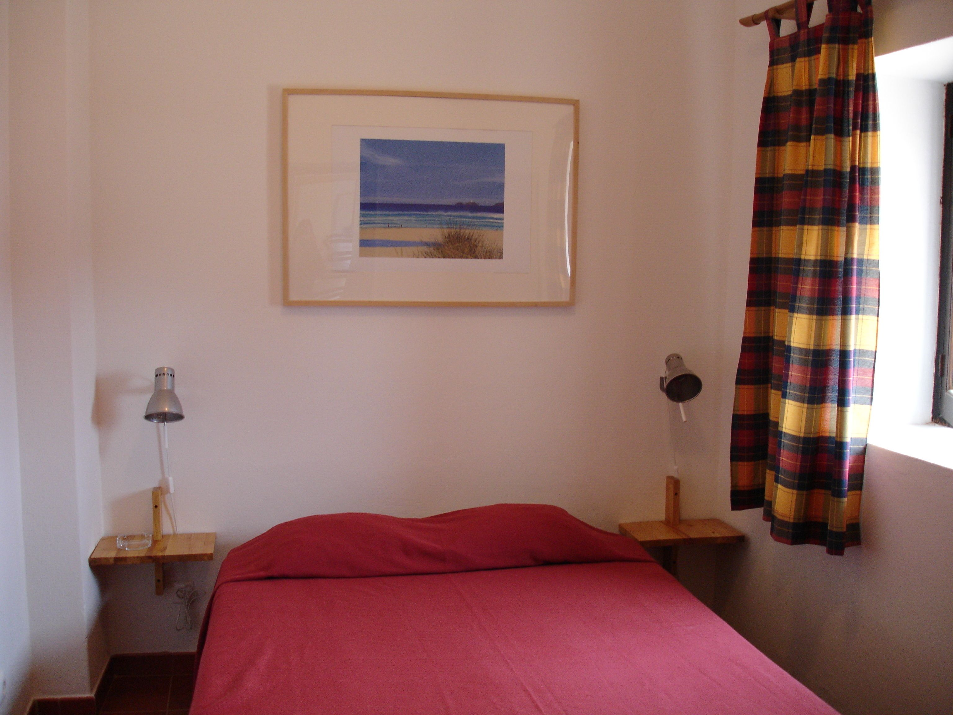 Une chambre à coucher avec un lit, un tableau encadré au mur, une lampe et un rideau à carreaux.