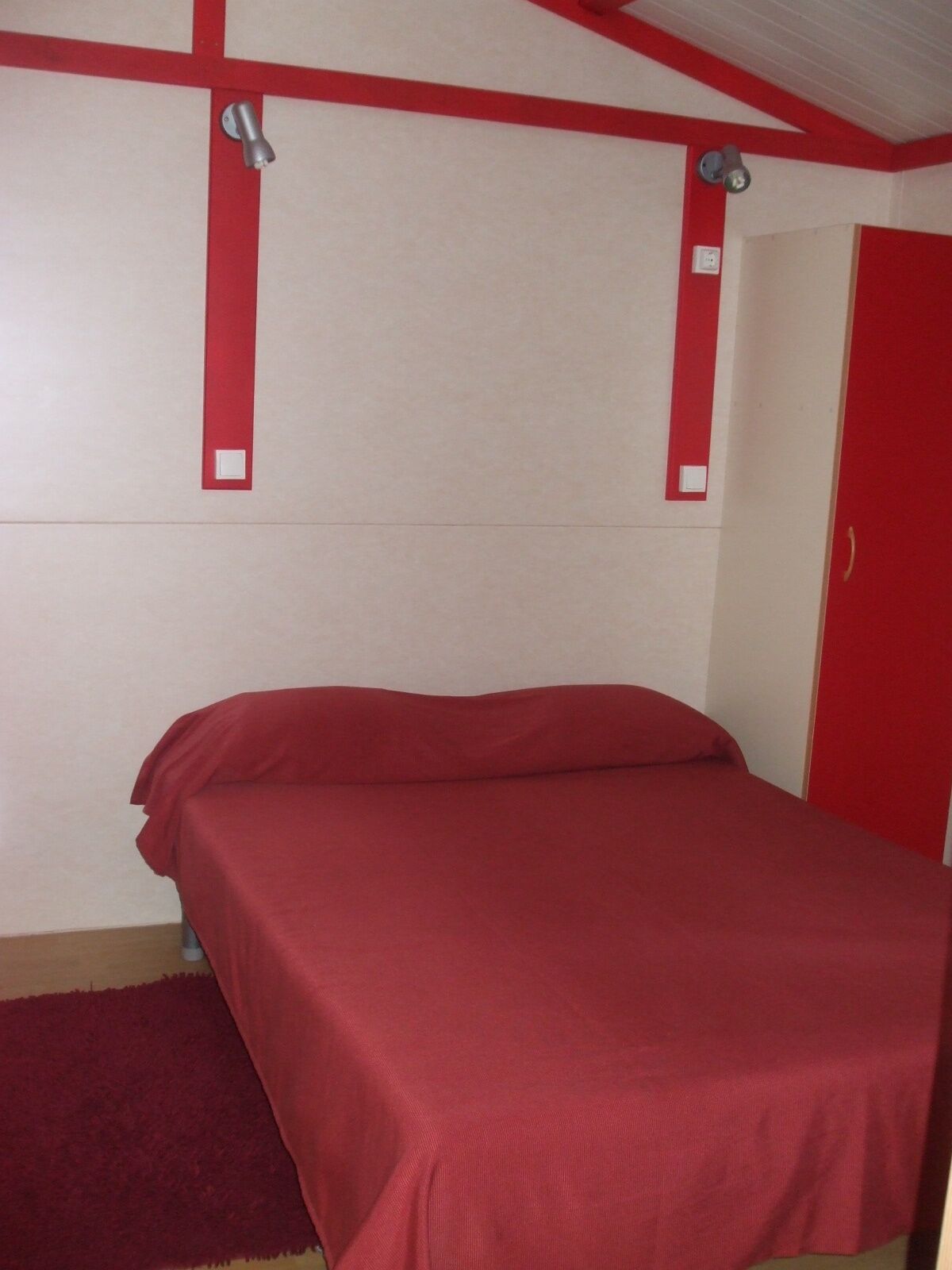 Uma cama de solteiro com um cobre-leito vermelho em um quarto com paredes brancas e detalhes vermelhos.