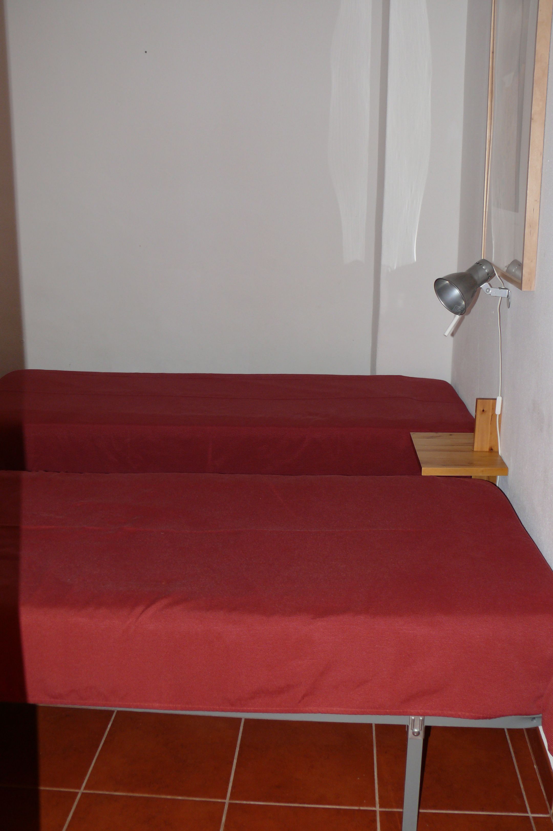 Una habitación con dos camas, cada una con una colcha roja, una mesita de noche de madera y una lámpara de pared.