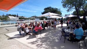 An outdoor patio area with tables and chairs, umbrellas, and a pool in the background. - Parque de Campismo Orbitur Valverde (Lagos)