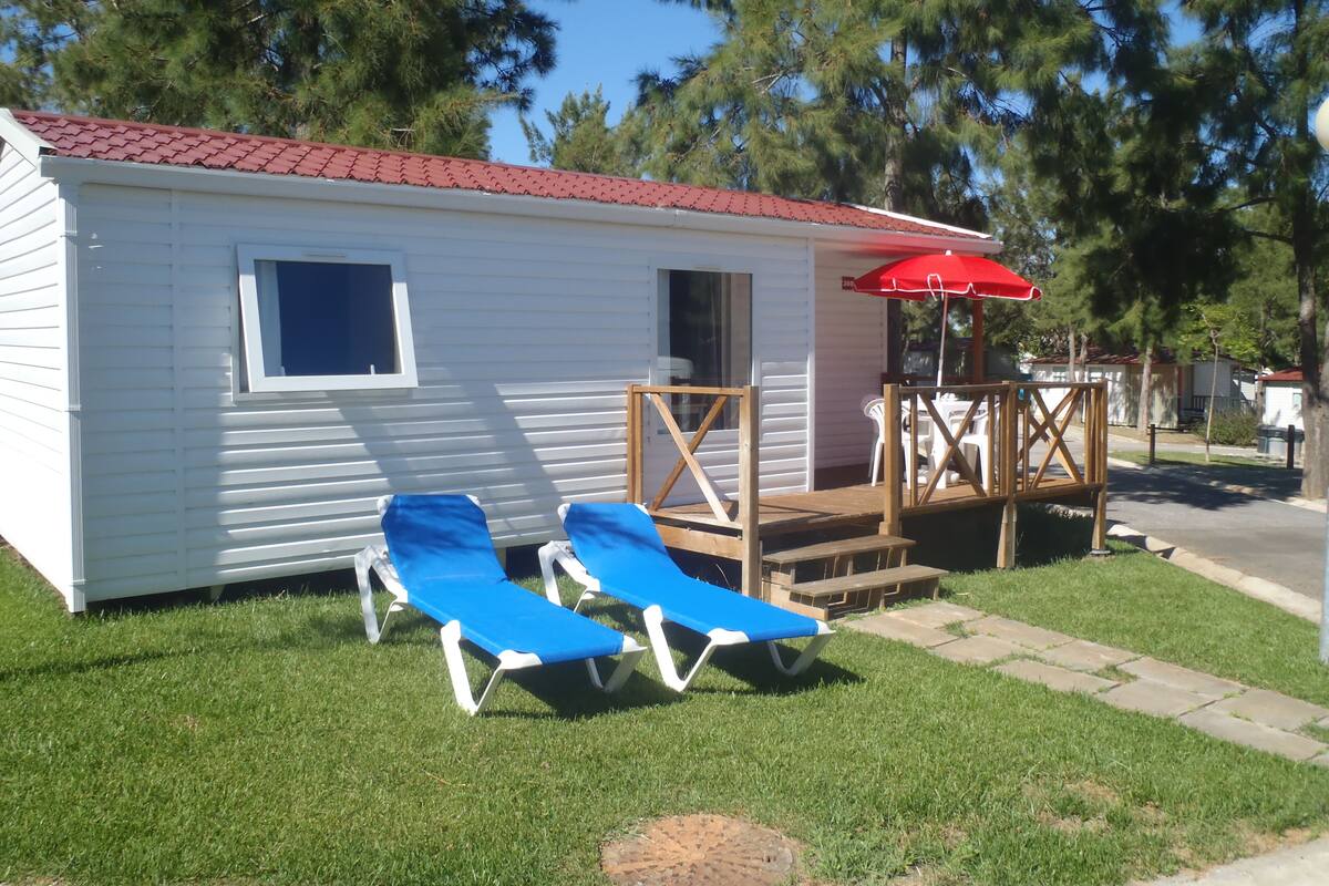 A small white cabin with a red roof, two blue lounge chairs, and a red umbrella on a grassy area.